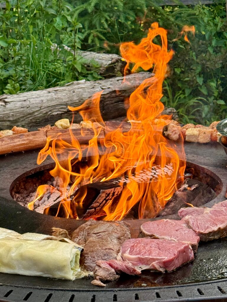 Auf dem Grill liegt Fleisch während in der Mitte des Grills das Feuer lodert.